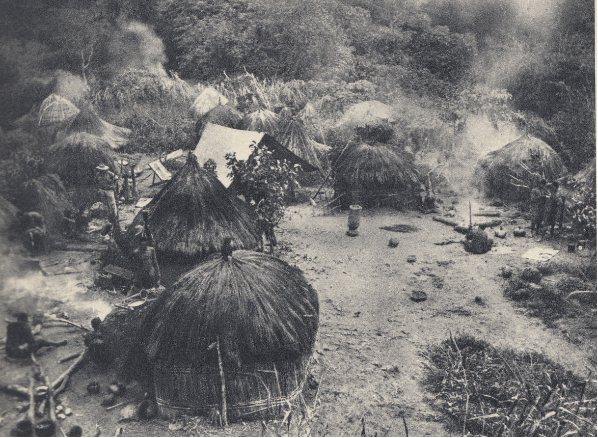 Historical photograph of a traditional Katanga village showing round thatched huts arranged in a clearing, with cooking fires and dense vegetation in the background.