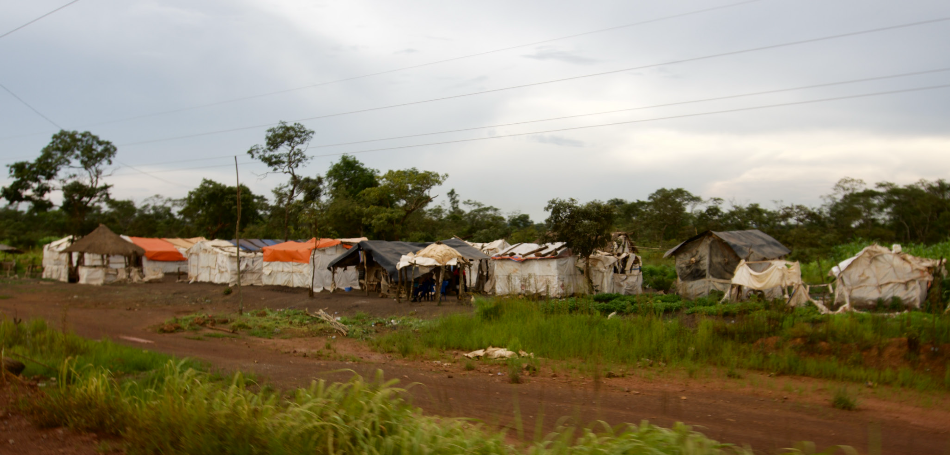 Makeshift tents and shelters of a temporary labour camp beside a mine access road, under a heavy grey sky.