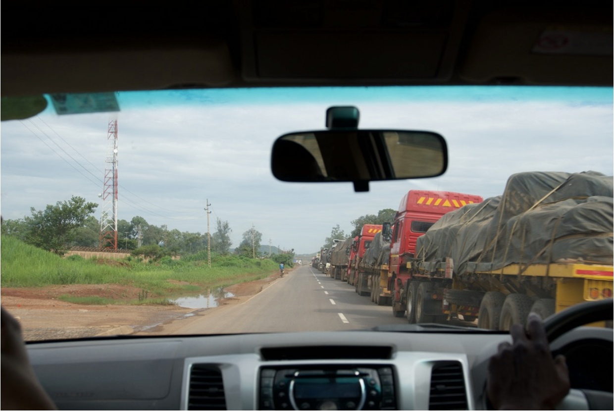 View from inside a vehicle on the road to Kolwezi: a long convoy of heavy mineral trucks stretches into the horizon, flanked by red-earth verges and green trees.